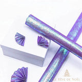 Rouleaux de papier cadeau violet irisé posés en croix sur une boîte cadeau blanche à rosaces