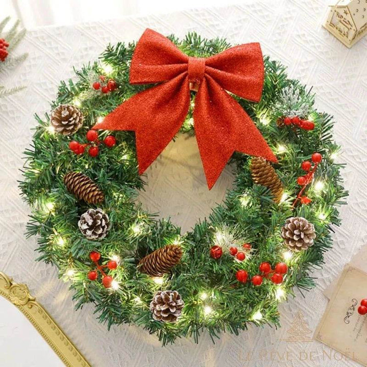 Couronne de Noël lumineuse posée à plat sur un tissu blanc, nœud rouge et pommes de pin