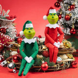 Deux figurines Grinch en peluche, une en tenue verte et une en tenue rouge, assises sur une couronne