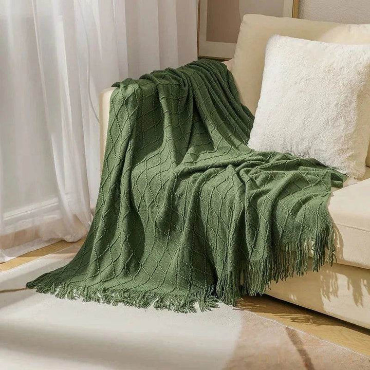 Plaid de Noël vert drapé sur un canapé crème, coussin blanc moelleux, rideaux voilage en fond