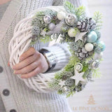 Couronne en osier blanc décorée de boules argentées, pommes de pin et branches, tenue en main