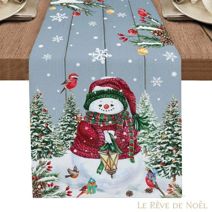 Même chemin de table bleu-gris, avec bonhomme de neige en bonnet et écharpe rouges tenant une lanterne, sapins enneigés