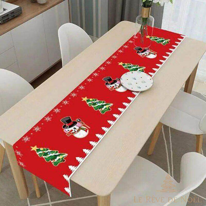 Chemin de table rouge à motif bonhommes de neige et sapins, posé en long sur une table en bois entre chaises blanches