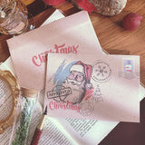 Deux enveloppes kraft roses illustrées d'un Père Noël, posées sur un livre ouvert avec une plume