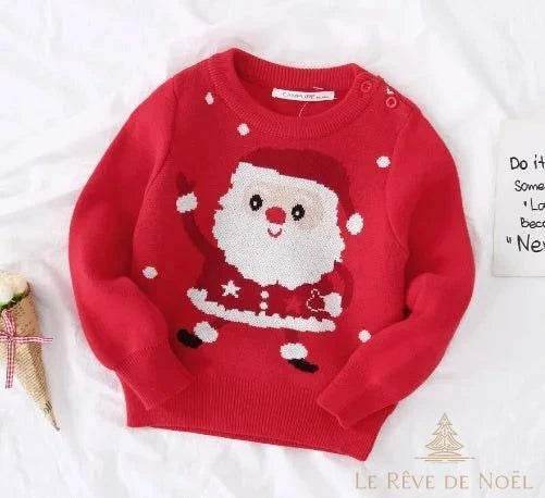Pull de Noël enfant rouge à motif Père Noël qui salue, posé à plat sur un lit avec décorations