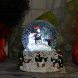 Boule à neige à pingouins allumée, posée près d'un sapin et de fleurs rouges, ambiance tamisée