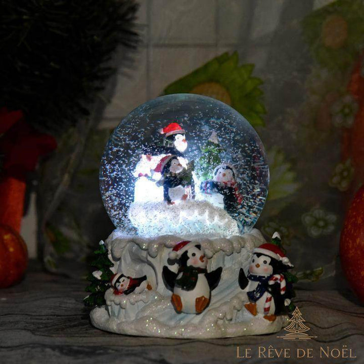 Boule à neige à pingouins allumée, posée près d'un sapin et de fleurs rouges, ambiance tamisée