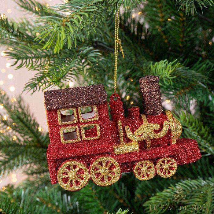 Décoration de Noël en forme de petit train rouge pailleté à roues dorées, suspendue à une branche de sapin