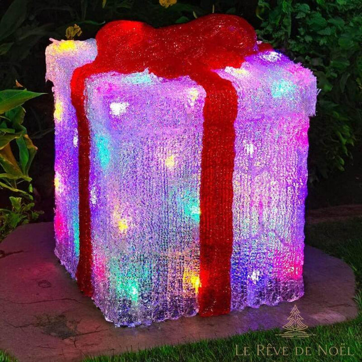 Grand cadeau lumineux en acrylique givré, ruban rouge, LED multicolores allumées, installé dehors la nuit