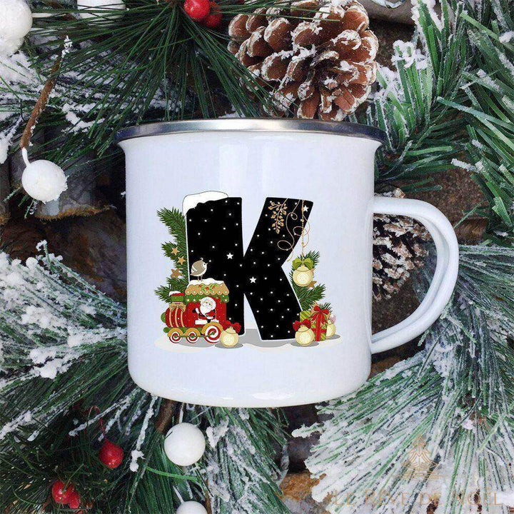 Mug émaillé blanc à lettre K noire décorée d'un petit train avec le Père Noël, branches de sapin givrées