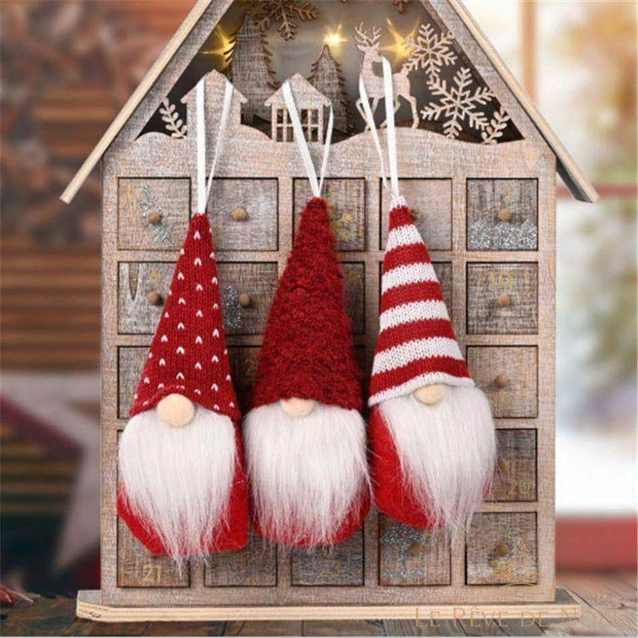 Trois lutins de Noël rouges à suspendre, bonnets à motifs différents et barbe blanche, devant une maison en bois