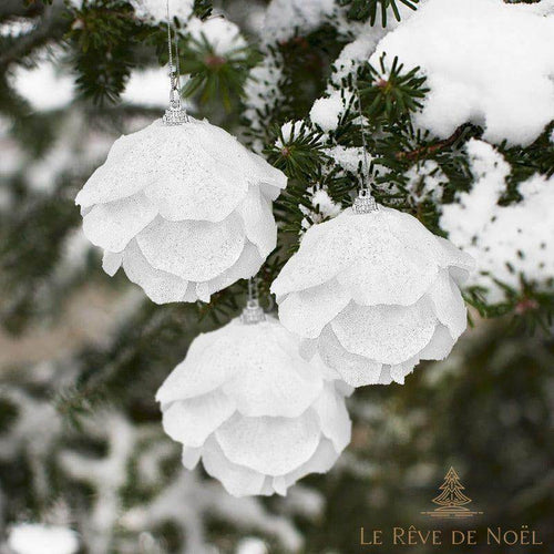 Trois boules de Noël blanches en forme de rose, suspendues à un sapin enneigé en extérieur
