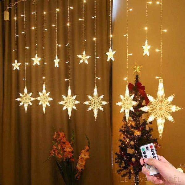 Rideau lumineux à étoiles suspendu devant un rideau marron, petit sapin décoré et télécommande en main